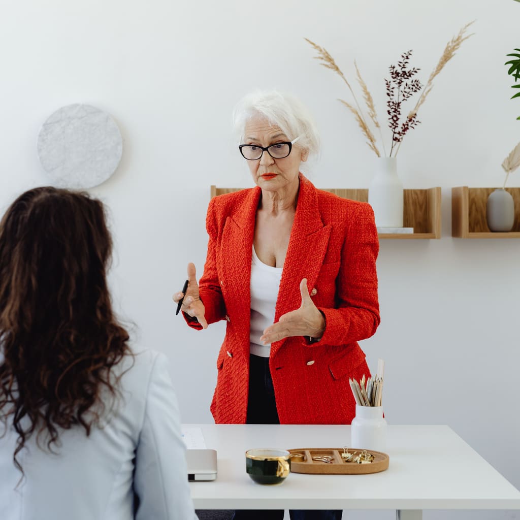 A professional in a red blazer gestures while speaking with a colleague across a desk.
