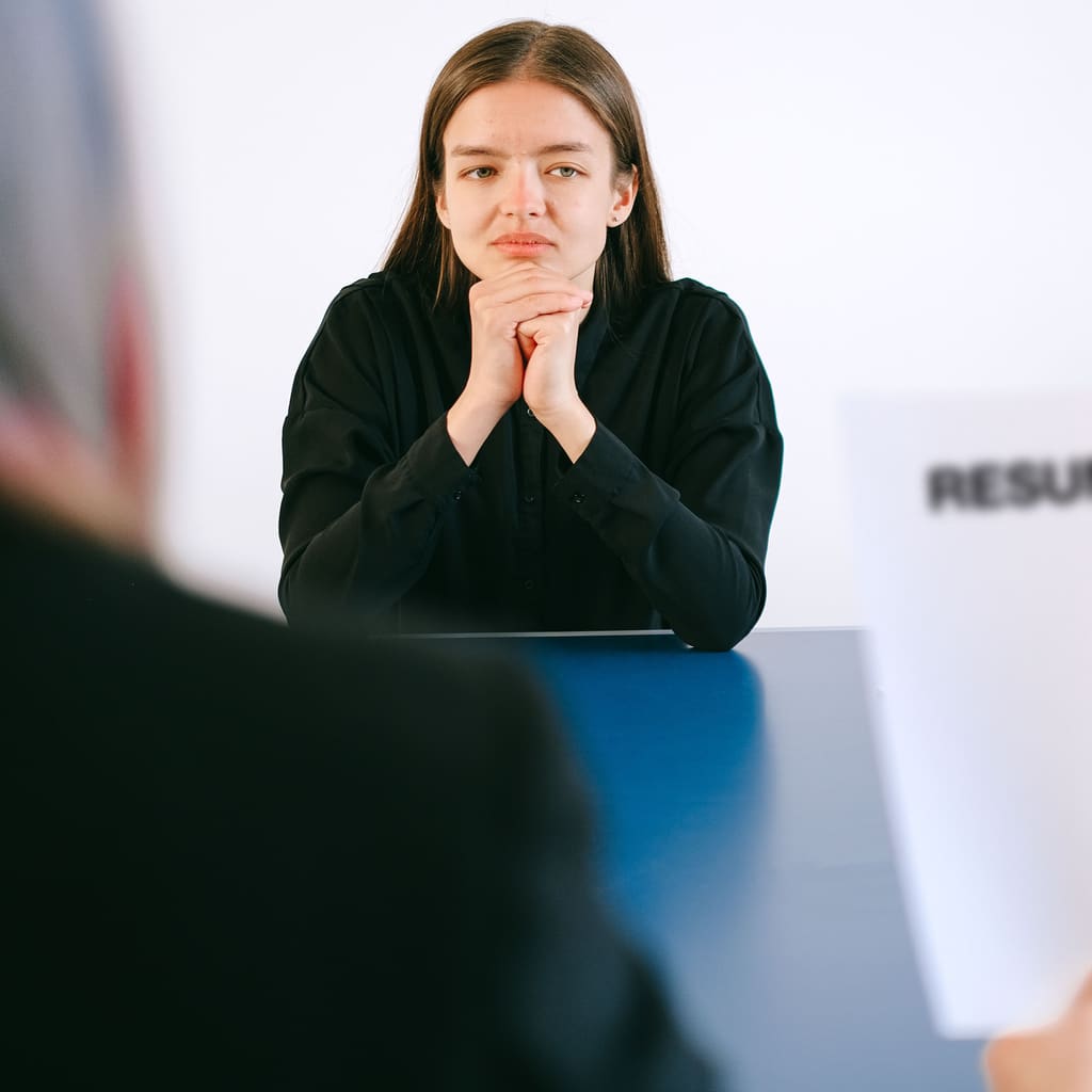A job candidate sits across the table from two interviewers, one of whom holds a printed resume.