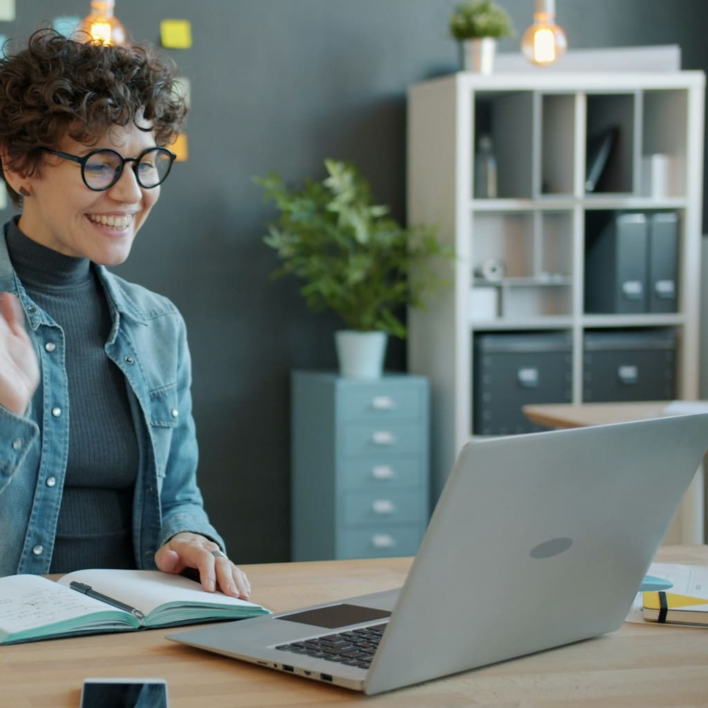 A smiling professional in glasses works at a desk with a laptop and notebook.