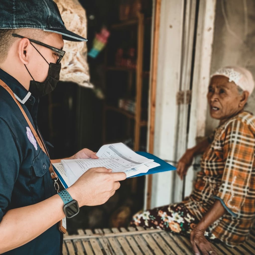 A social worker with a clipboard talks with an elderly resident at their doorway.