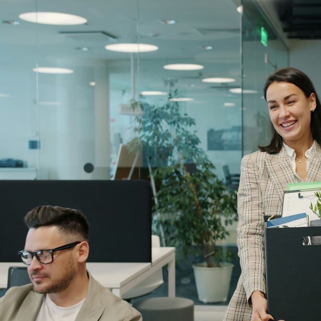 A smiling employee carries a box of belongings through an office.